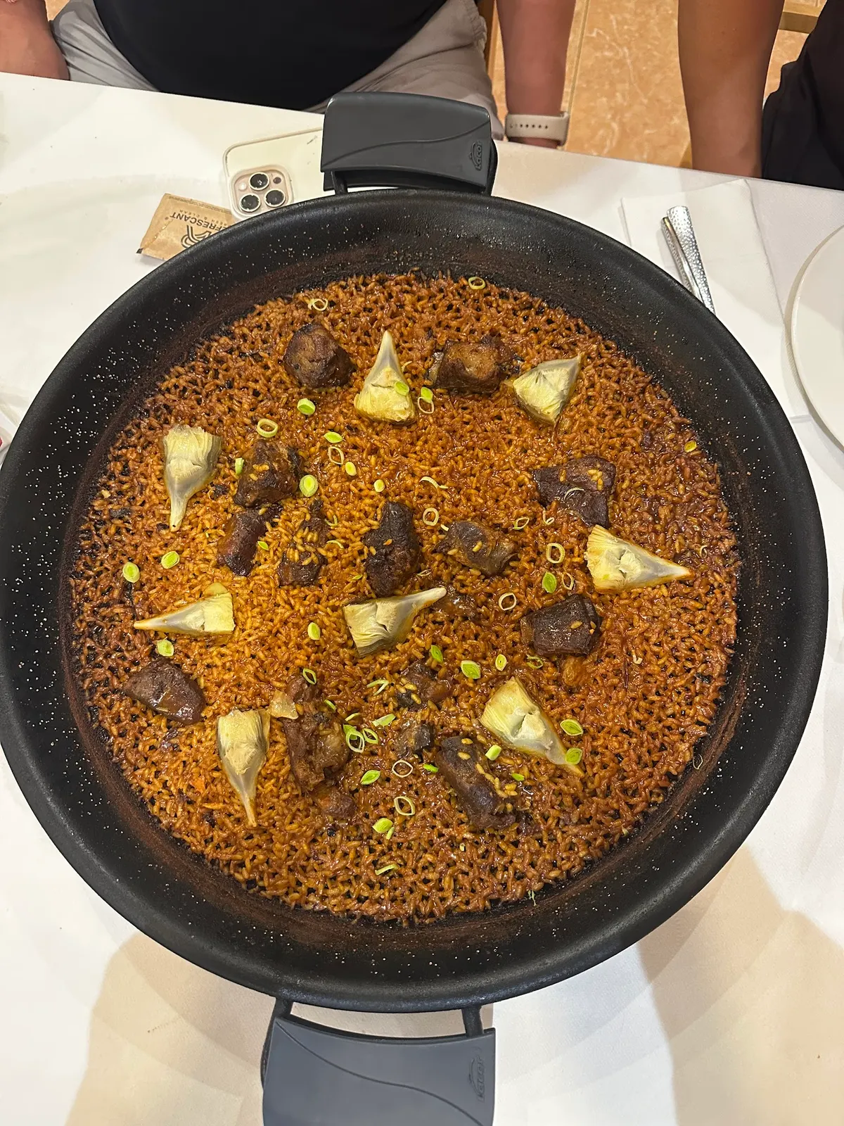 Paella vista desde arriba con arroz tostado, trozos de carne, alcachofas y ajos tiernos, sobre una mesa con mantel blanco