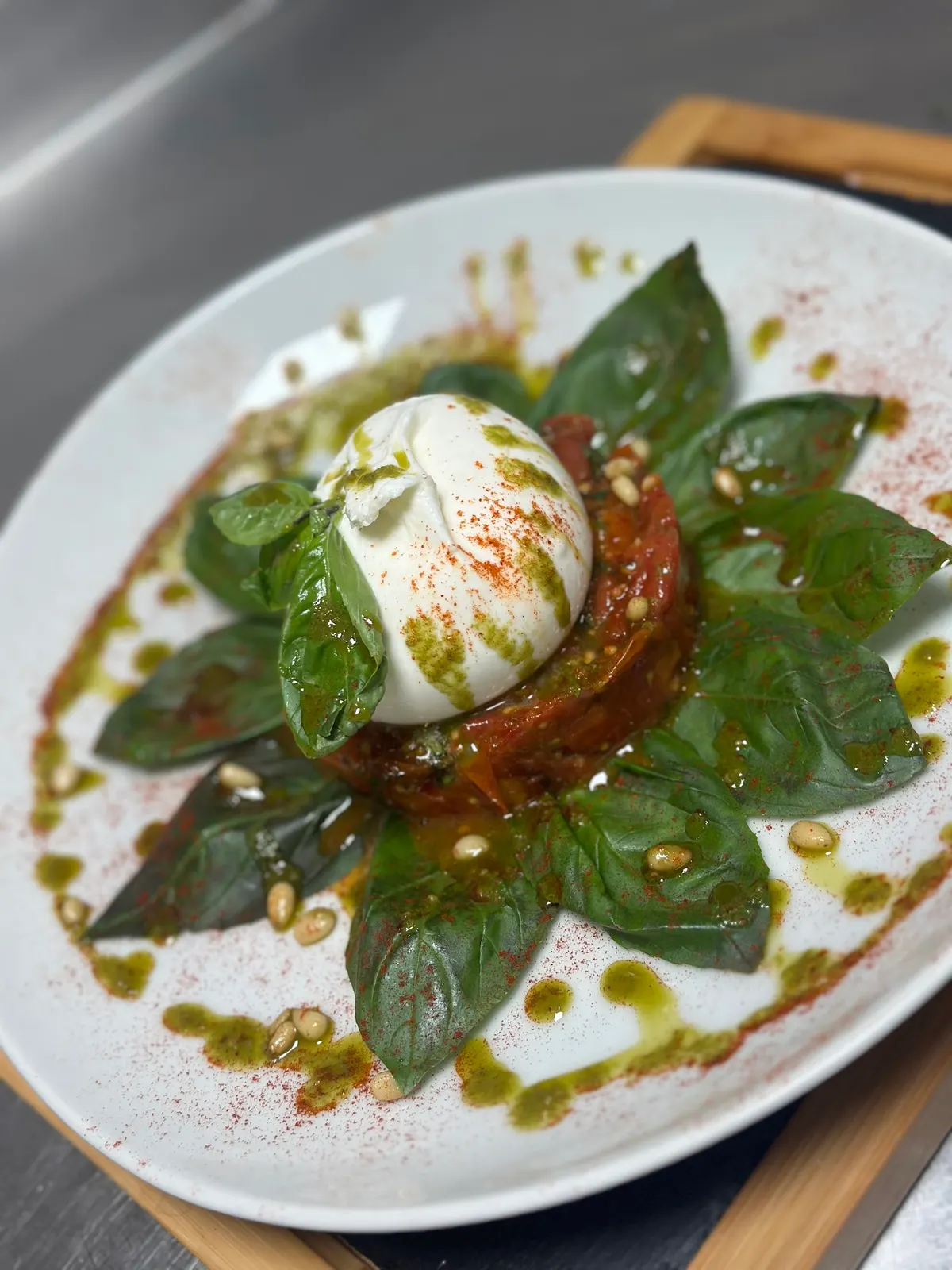 Burrata sobre tomate y hojas de albahaca dispuestas en estrella, con piñones, pesto y pimentón en plato blanco