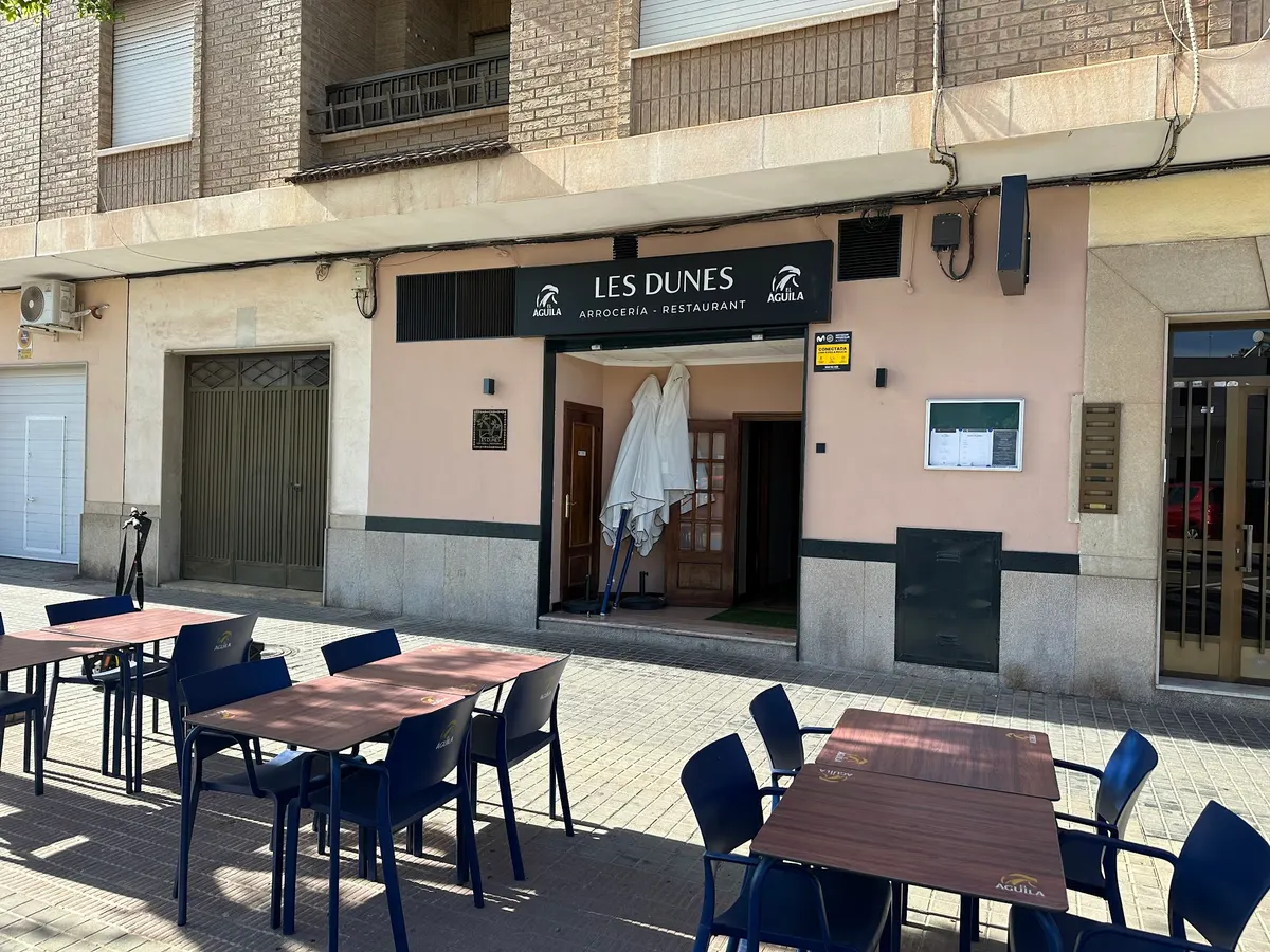 Fachada de Les Dunes en la avenida Jaume I con su rótulo negro y la terraza de mesas de madera y sillas azules al sol