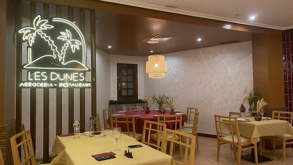 Sala de Les Dunes con el rótulo de neón de las palmeras y «Les Dunes, arrocería - restaurant», mesas con mantel y sillas de madera clara